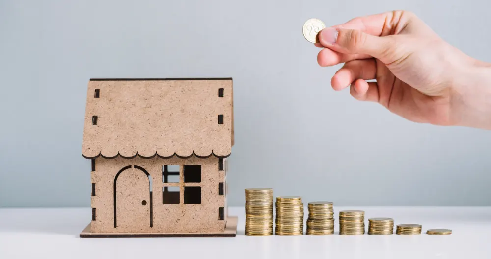 increasing piles of coins in front of a cardboard house showing the concept of How To Secure Your Home On A Budget